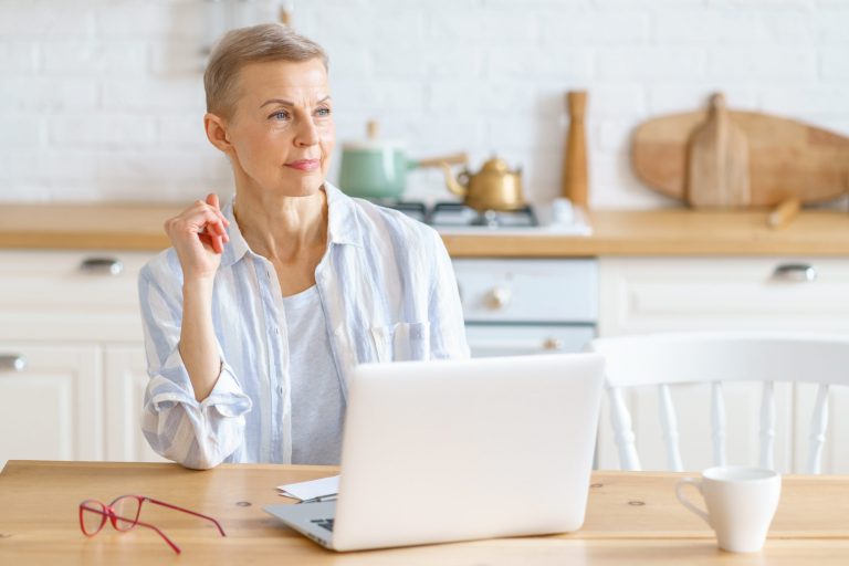 Happy dreaming senior woman in casual clothes looking aside and thinking while working or surfing internet on laptop, happy elderly 60s woman enjoying morning with coffee in light modern kitchen